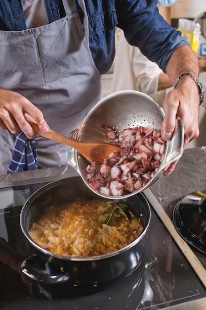Chef masculin méconnaissable debout à la cuisinière et ajoutant du poulpe coupé à un délicieux plat avec des légumes dans une casserole pendant la cuisson dans la cuisine à la maison
