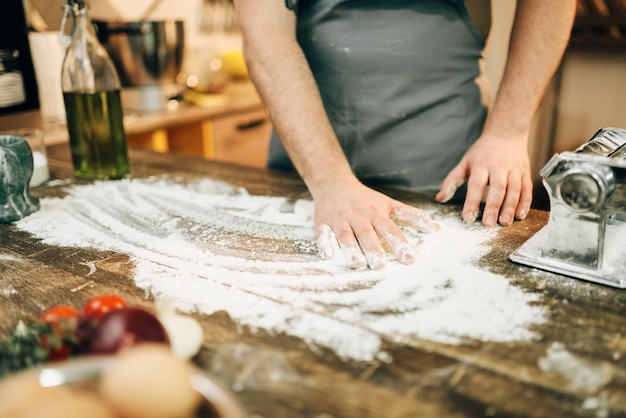 Chef masculin en tablier, farine, œufs et machine à pâtes sur table en bois. Ingrédients pour spaghetti maison