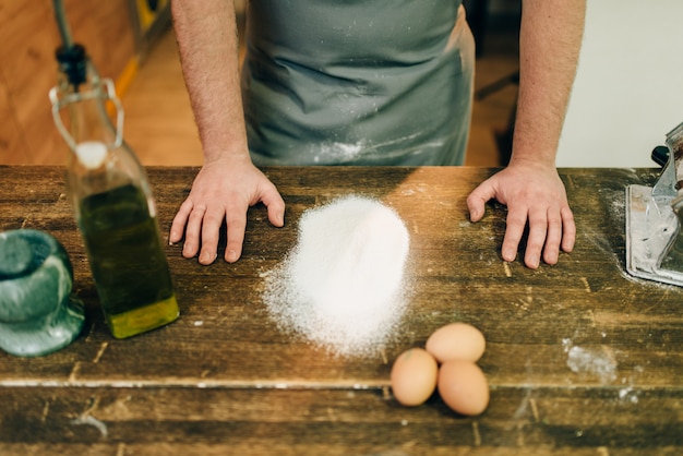 Chef masculin en tablier, farine et œufs sur table en bois. Cuisson des pâtes maison