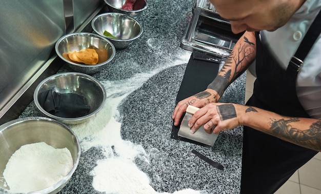 chef masculin avec des tatouages sur ses mains coupant la pâte pour les pâtes faites maison sur la table de cuisine