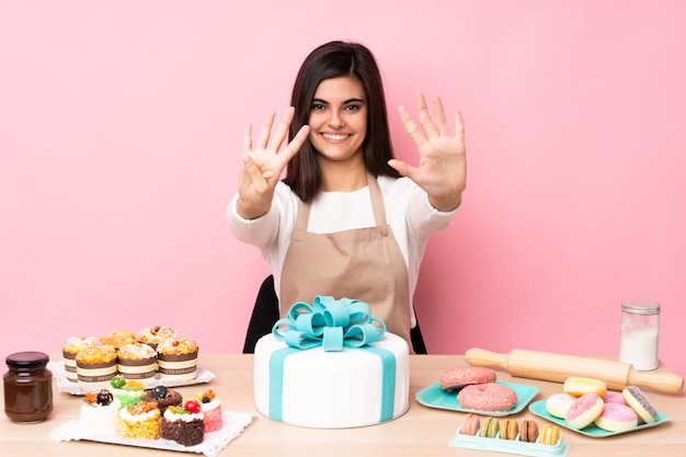 Chef pâtissier avec un gros gâteau dans une table sur un mur rose isolé comptant neuf avec les doigts