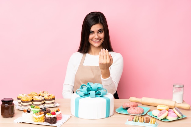 Chef pâtissier avec un gros gâteau dans une table sur un mur rose isolé invitant à venir avec la main. Heureux que tu sois venu