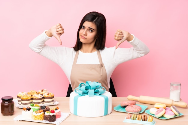 Chef pâtissier avec un gros gâteau dans une table sur un mur rose isolé montrant le pouce vers le bas