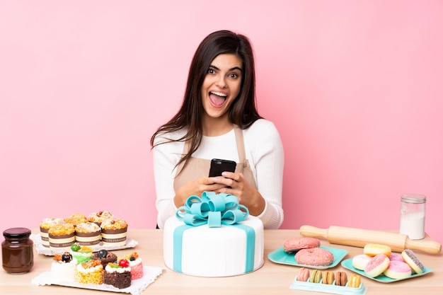 Chef pâtissier avec un gros gâteau dans une table sur un mur rose isolé surpris et en envoyant un message