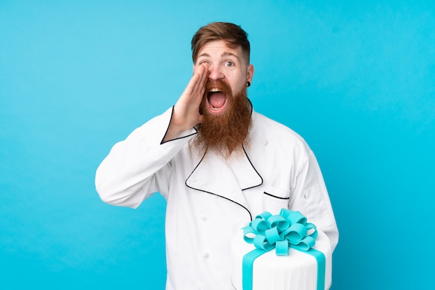 Chef pâtissier rousse avec longue barbe tenant un gros gâteau sur le mur bleu isolé criant avec la bouche grande ouverte