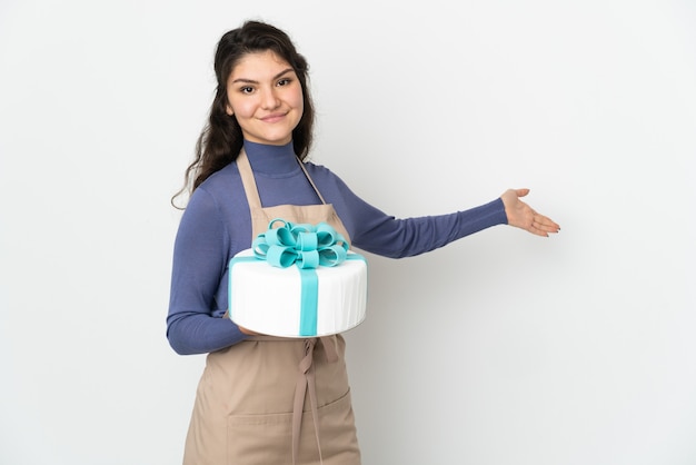 Chef pâtissier russe tenant un gros gâteau isolé sur un mur blanc étendant les mains sur le côté pour inviter à venir