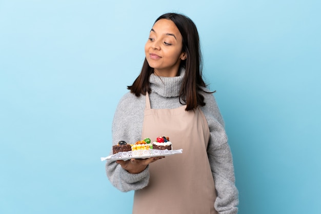 Chef pâtissier tenant un gros gâteau sur bleu isolé ayant des doutes tout en regardant côté