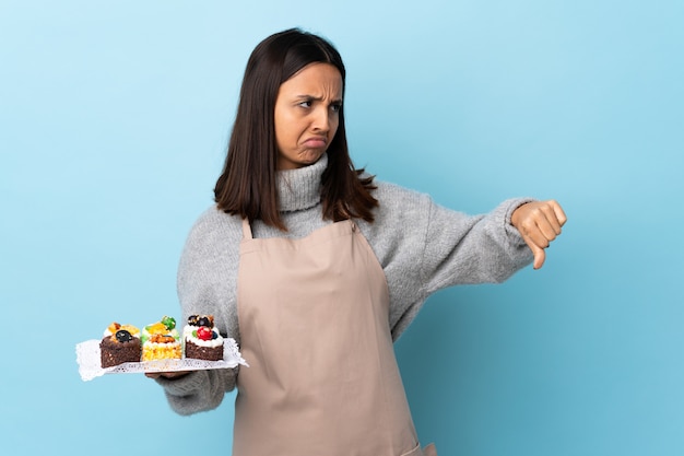 Photo chef pâtissier tenant un gros gâteau sur bleu isolé montrant le pouce vers le bas avec une expression négative.