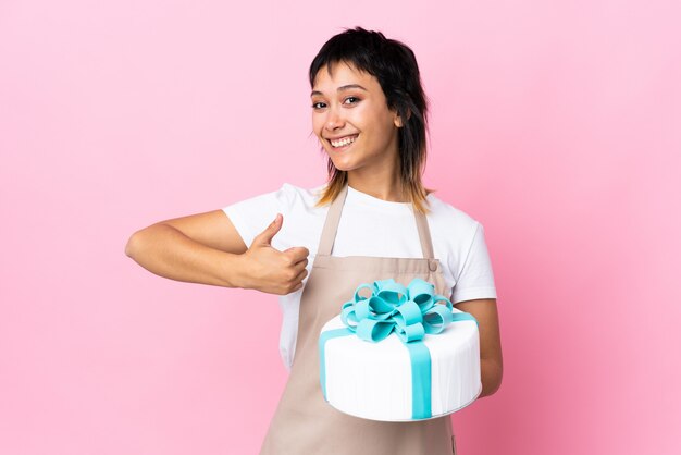 Chef pâtissier tenant un gros gâteau sur un espace rose isolé donnant un geste du pouce levé