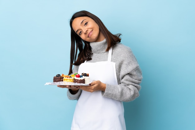 Chef pâtissier tenant un gros gâteau sur un mur bleu isolé maintient la paume ensemble. La personne demande quelque chose.