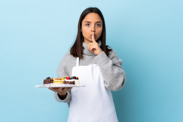 Chef pâtissier tenant un gros gâteau sur le mur bleu pointant vers le côté et faisant un geste de silence.