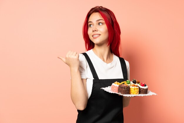 Chef pâtissier tenant un muffins isolé sur un mur rose pointant vers le côté pour présenter un produit