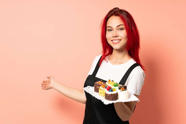 Chef pâtissier tenant un muffins isolé sur un mur rose tendant les mains sur le côté pour inviter à venir