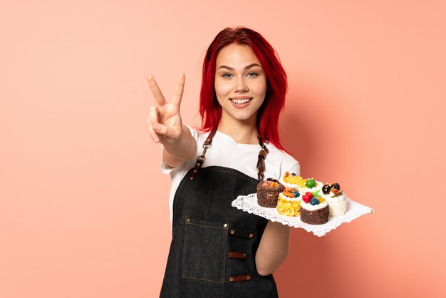 Chef pâtissier tenant un muffins isolé sur rose souriant et montrant le signe de la victoire