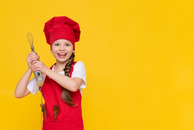 Chef petite fille tient un mixeur pour fouetter Un enfant dans un bonnet rouge et un tablier sur un fond jaune isolé Une place pour la publicité Copier l'espace