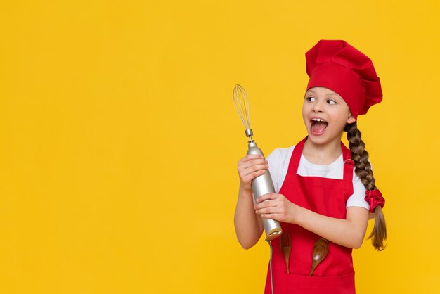 Chef petite fille tient un mixeur pour fouetter Un enfant dans un bonnet rouge et un tablier sur un fond jaune isolé Une place pour la publicité Copier l'espace