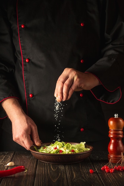 Le chef professionnel arrose le sel à la salade des légumes frais sur la table en bois