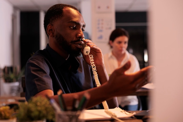 Le chef de projet discute du rapport de l'entreprise avec un collègue sur un téléphone fixe, l'employé répond à l'appel tardif au bureau. Homme afro-américain ayant une communication téléphonique dans un espace de coworking