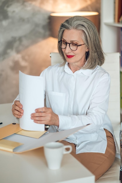 Chef de projet. Femme aux cheveux gris assise à la table et travaillant avec la documentation du projet