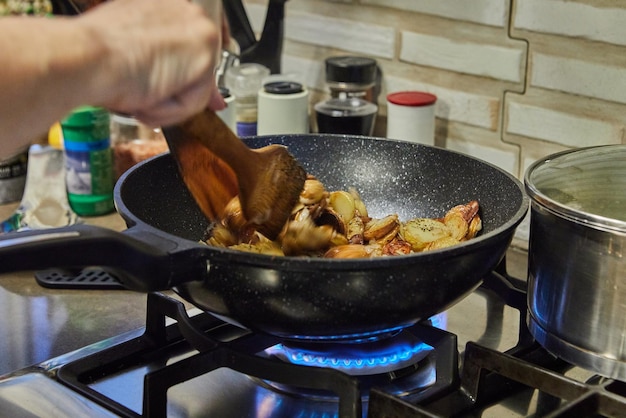 Le chef remue des tranches de pommes de terre avec des champignons dans une poêle à frire sur une cuisinière à gaz