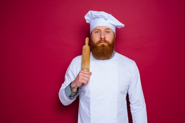 Le chef sérieux avec la barbe et le chef de tablier rouge tient le rouleau à pâtisserie en bois