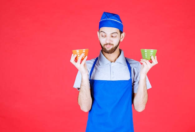 Chef en tablier bleu tenant des tasses en céramique jaune et verte dans les deux mains.