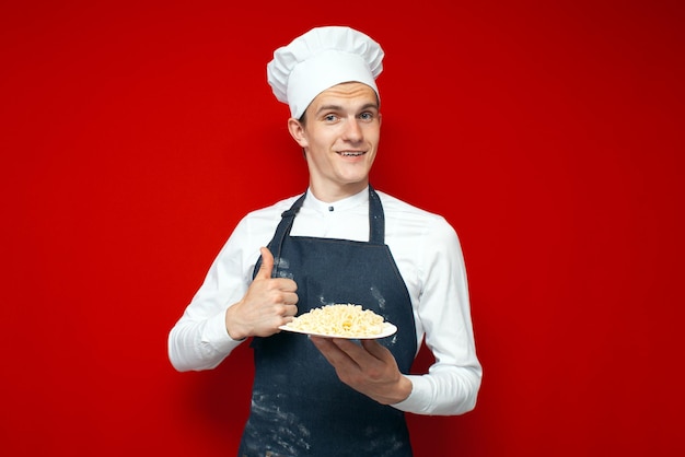 Le chef tient un plat sur fond isolé rouge et montre comme un ouvrier de cuisine dans des pâtes cuites uniformes