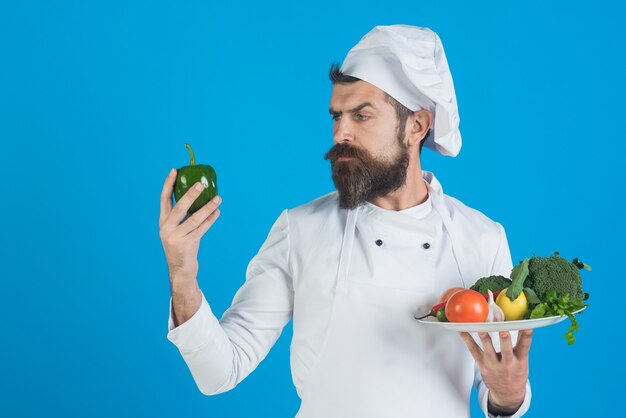 Le chef avec le visage sérieux dans l'uniforme blanc tient des légumes sur le concept vert d'épicerie de plat faisant cuire et