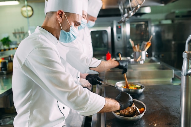 Des chefs portant des masques de protection et des gants préparent la nourriture dans la cuisine d'un restaurant ou d'un hôtel.