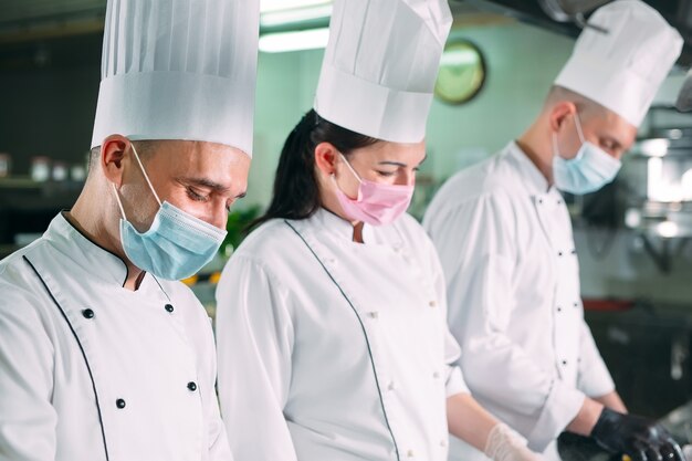 Les chefs portant des masques de protection et des gants préparent la nourriture dans la cuisine d'un restaurant ou d'un hôtel.