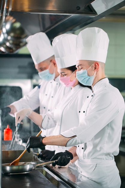 Les chefs portant des masques de protection et des gants préparent la nourriture dans la cuisine d'un restaurant ou d'un hôtel.