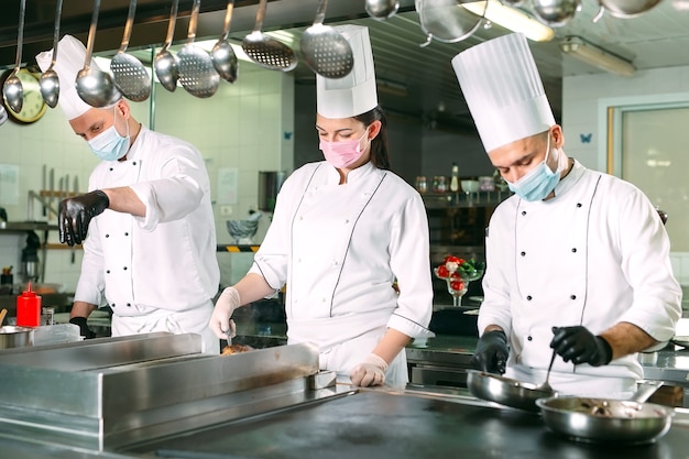Les chefs portant des masques de protection et des gants préparent la nourriture dans la cuisine d'un restaurant ou d'un hôtel.