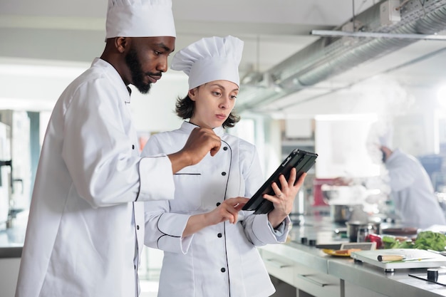 Des chefs professionnels avec un appareil à écran tactile sans fil préparant des ingrédients pour un concours culinaire organisé au restaurant. Experts en cuisine gastronomique avec tablette moderne à la recherche d'idées de plats pour le dîner sur Internet.