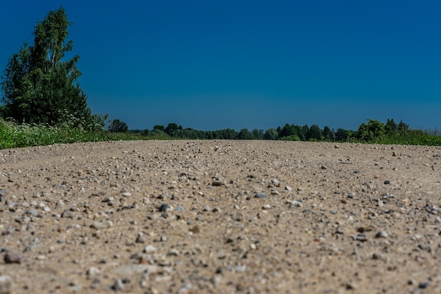 Un chemin de terre déserte de sable et de petites pierres, entouré d'herbe et d'arbres, contre un ciel bleu.