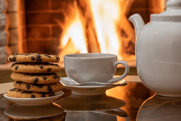 Cheminée confortable, théière et tasse à thé, biscuits sur une table., reflet d'un feu sur une table en verre.