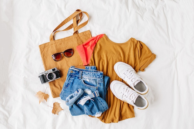 Chemise beige, jean bleu, baskets, sac écologique et appareil photo sur lit sur drap blanc. Tenue d'automne élégante pour femmes. Collage de vêtements à la mode. Mise à plat, vue de dessus.