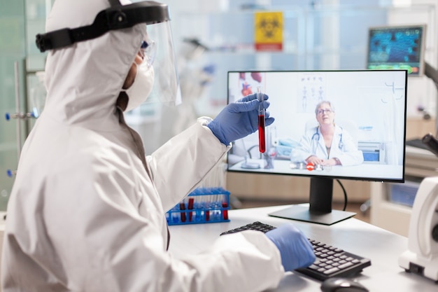 Chercheur de laboratoire discutant avec un appel vidéo vêtu d'un costume en ppe montrant un tube à essai. Scientifique montrant un échantillon de sang lors d'une discussion en ligne, recherchant un traitement contre le virus covid19.