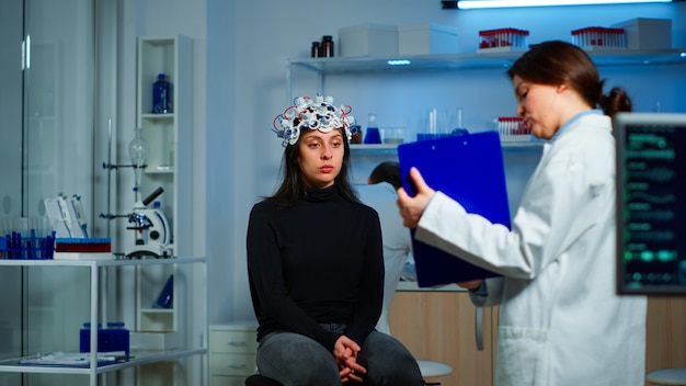 Chercheur médical pointant sur le presse-papiers expliquant le traitement contre les maladies du cerveau au patient avec un casque eeg. Femme assise dans un laboratoire scientifique neurologique traitant les dysfonctionnements du système nerveux