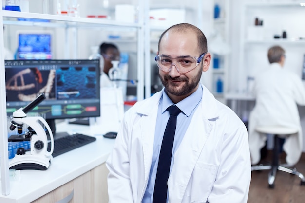 Chercheur pharmaceutique assis sur un tabouret regardant la caméra.Sérieux expert en génétique en laboratoire avec une technologie moderne pour les investigations médicales avec un assistant africain en arrière-plan.