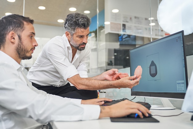 Des chercheurs masculins barbus concentrés examinent un modèle de masque facial imprimé tout en travaillant sur un ordinateur avec une illustration 3D d'un respirateur dans un bureau contemporain