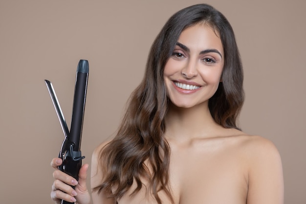 Cheveux, appareil. Jolie jeune femme souriante aux épaules nues avec fer à friser de bonne humeur