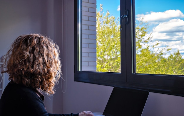 Cheveux blonds bouclés femme adulte travaillant sur son ordinateur devant la fenêtre.