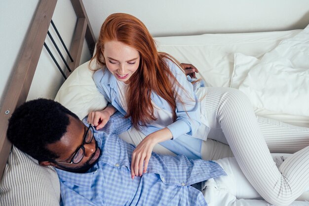 Cheveux roux femme européenne et beau mâle afro-africain ensemble étreignant allongé dans la chambre à la maison appartement confortable