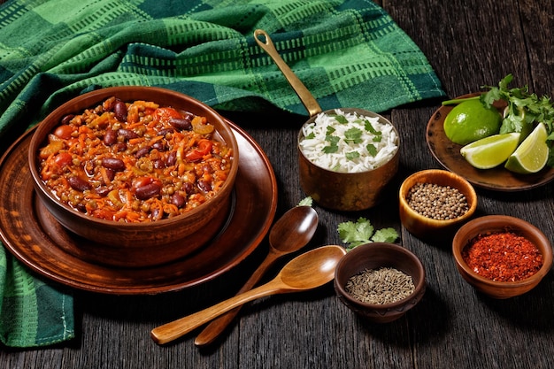 Chili végétarien aux haricots rouges et lentilles dans un bol en terre cuite sur une table en bois sombre avec du riz et des épices, cuisine américaine