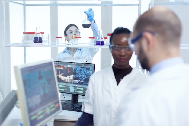 Photo chimiste expert examinant un liquide dans un récipient en verre. équipe multiethnique de chercheurs en médecine travaillant ensemble dans un laboratoire stérile portant des lunettes et des gants de protection.