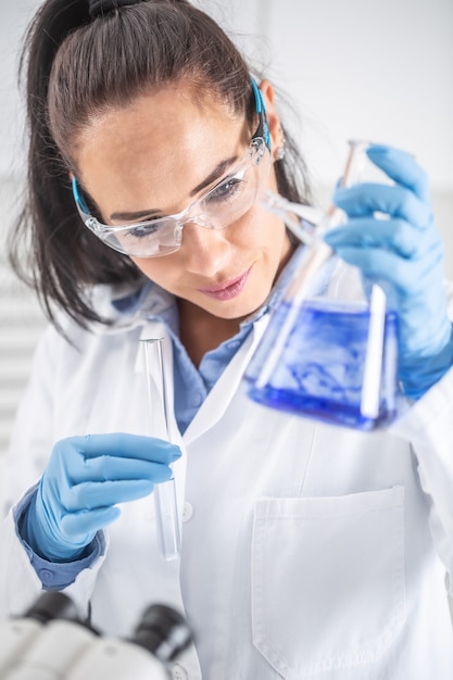 Une chimiste portant des lunettes de protection sourit en tenant un tube à essai, mélangeant un liquide bleu à l'intérieur de la fiole conique. Concept de coronavirus COVID-19.