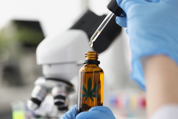 Chimiste scientifique dégoulinant d'huile de marijuana de la pipette dans le pot sur fond de microscope