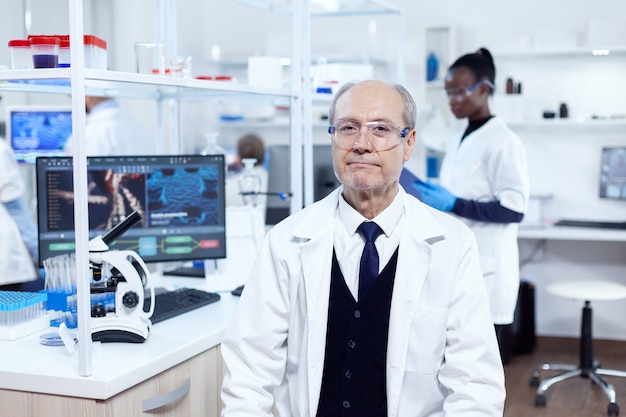 Chimiste senior masculin regardant la caméra portant des lunettes de protection. Scientifique âgé portant une blouse de laboratoire travaillant à la mise au point d'un nouveau vaccin médical avec un assistant africain en arrière-plan.