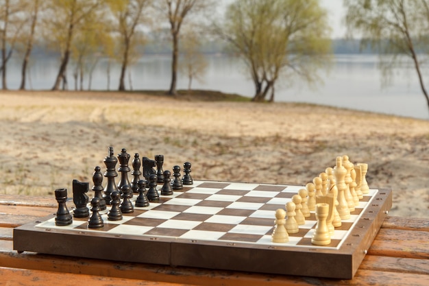 Échiquier avec pièces d'échecs sur banc en bois avec fond de remblai de rivière. Jeu d'échecs avec des pièces d'échecs en bois