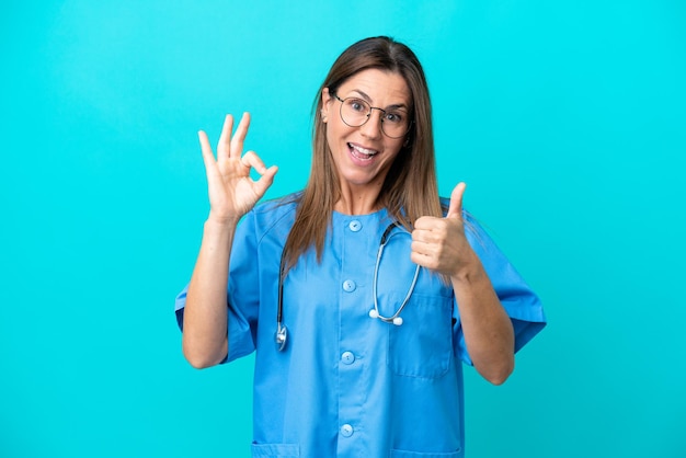 Chirurgien d'âge moyen femme isolée sur fond bleu montrant le signe ok et le geste du pouce vers le haut